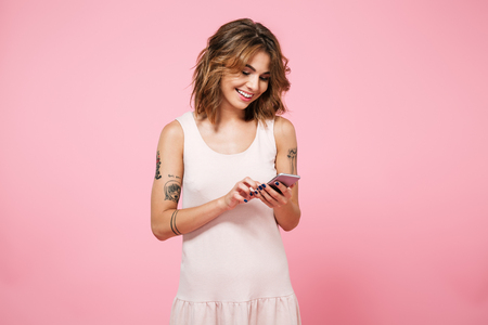 Portrait of a pretty smiling girl in summer clothes using mobile phone while standing isolated over pink backgroundの写真素材