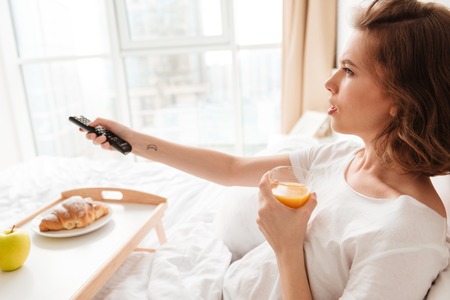 Side view picture of serious young lady sitting indoors at home with croissant and juice. Looking aside watch tv holding remote control.の写真素材