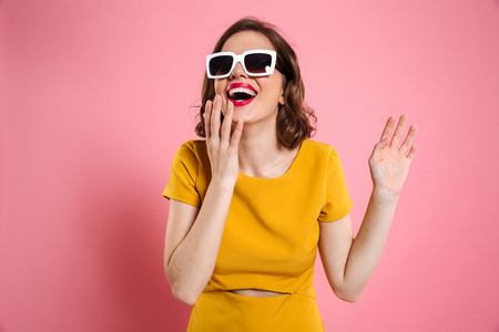 Portrait of a happy young woman in sunglasses and dress standing and laughing isolated over pink backgroundの写真素材