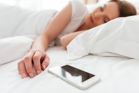 Photo of sleeping young woman lies in bed with eyes closed near mobile phone.の写真素材