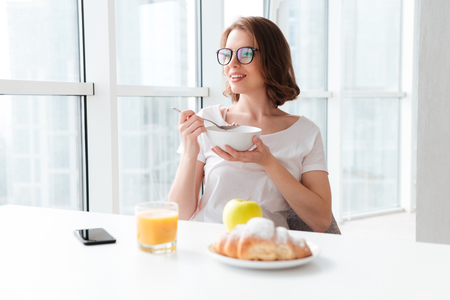 Picture image of cheerful young pretty woman sitting indoors at the table with juice and croissant eating corn flakes. Looking aside.の写真素材