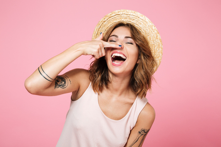 Portrait of a smiling funny girl in summer hat holding finger at her nose isolated over pink backgroundの写真素材