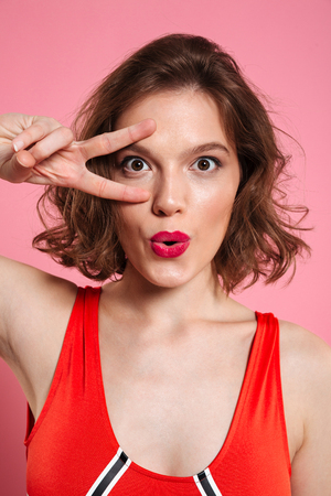 Close-up portrait of beautiful young woman with red lips showing peace gesture, looking at camera, isolated on pink backgroundの写真素材