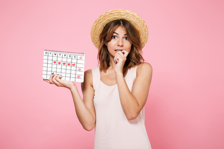 Portrait of embarrassed ?ute girl in summer hat holding calendar with drawn hearts and looking at camera isolated over pink backgroundの写真素材