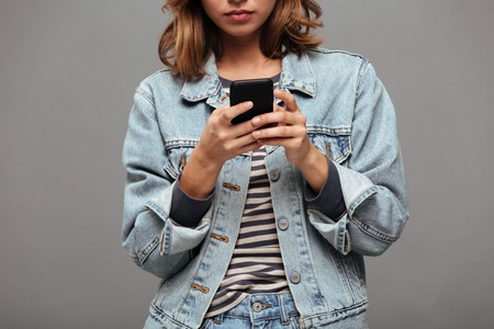 Cropped image of a serious young teenage girl dressed in denim jacket texting on mobile phone...