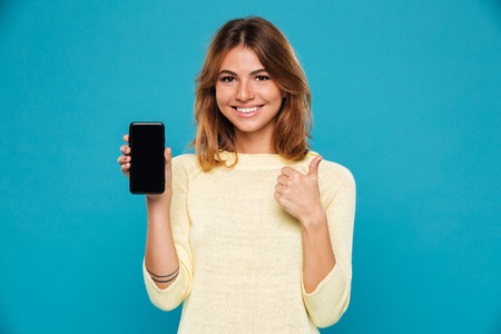 Smiling woman in sweater showing blank smartphone screen and thumb up while looking at the camera...