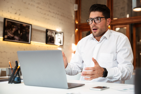 Close-up photo of amazed young businessman in glasses and white shirt looking at laptop screen in officeの写真素材