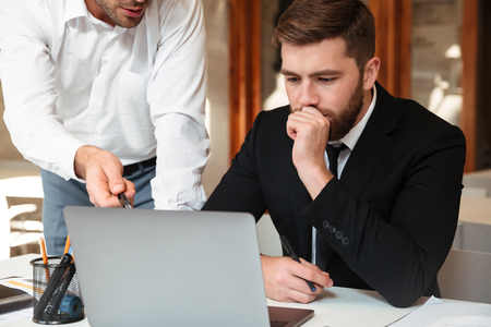 Close-up photo of two young man sitting in office and discussing business using laptopの写真素材