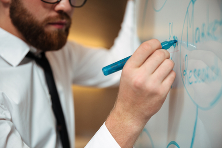 Close up portrait of a young bearded businessman in eyeglasses writing on a blackboard indoorsの写真素材