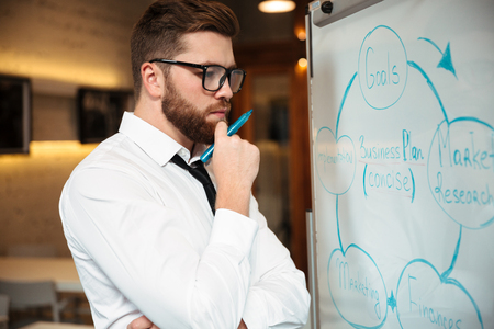 Portrait of a thoughtful bearded businessman holding hand with a pen at his chin and looking at blakboard indoorsの写真素材
