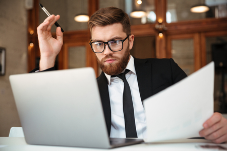 Young concentrated bearded businessman in formalwear looking at laptop screen holding papers while sitting at workplaceの写真素材