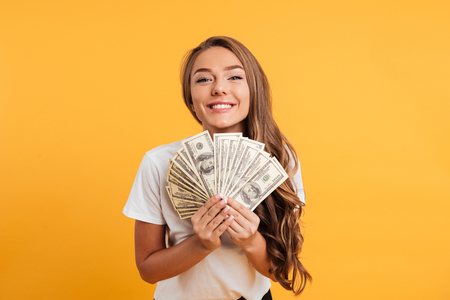 Portrait of a happy satisfied girl holding bunch of money banknotes and looking at camera isolated over yellow backgroundの写真素材
