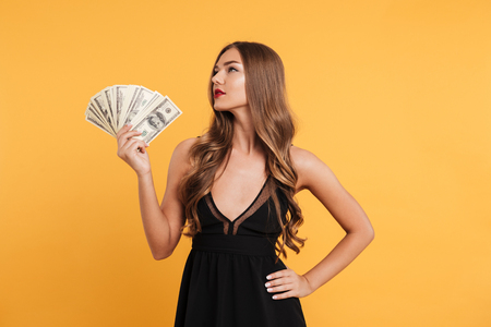 Portrait of a young attractive girl with long blond hair holding money banknotes and looking away isolated over yellow backgroundの写真素材