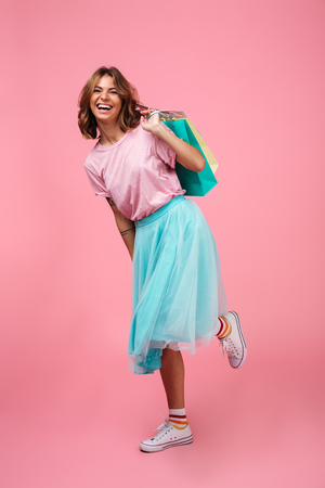 Full length portrait of a happy smiling girl in bright colorful clothes holding shopping bags while standing and looking at camera isolated over pink backgroundの写真素材
