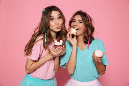 Two charming girls in colorful clothes enjoying tasty cupcakes, looking at camera isolated on pink backgroundの写真素材