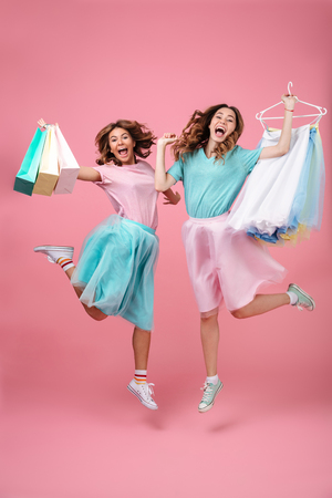 Full length portrait of two excited joyful girls dressed in bright colorful clothes holding shopping bags and jumping isolated over pink backgroundの写真素材