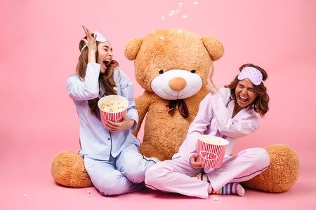 Two happy pretty girls dressed in pajamas having fun while sitting sitting with a big teddy bear and eating popcorn isolated over pink backgroundの写真素材