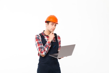 Portrait of a pensive young male builder working on laptop computer while standing and looking up isolated over white backgroundの写真素材