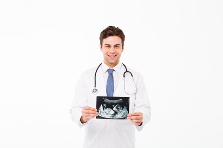 Portrait of a cheerful young male doctor with stethoscope dressed in uniform holding x-ray photograph and looking at camera isolated over white backgroundの写真素材
