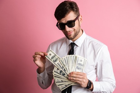 Portrait of a smiling confident man in sunglasses and white shirt counting money banknotes isolated...
