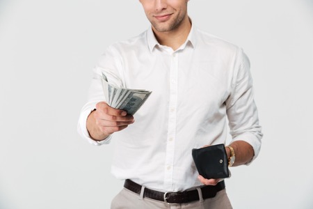Cropped image of a smiling man holding wallet and giving money banknotes isolated over white...