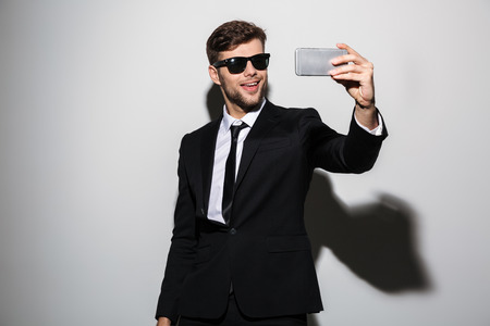 Cool bearded guy in black suit and suglasses taking selfie on mobile phone, isolated over white backgroundの写真素材