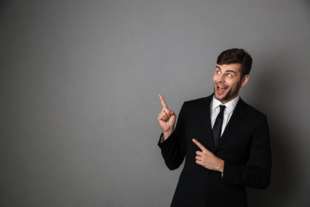 Overjoyed brunette man in classic suit pointing with two fingers, looking aside, isolated on gray backgroundの写真素材