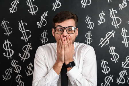 Close-up portrait of shocked man in glasses covering his mouth, looking at camera, isolated on black chalkboard with money symbolの写真素材