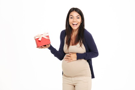 Portait of a happy young pregnant woman showing gift box and touching her belly isolated over white backgroundの写真素材