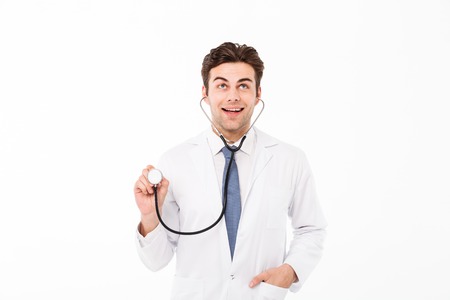 Portrait of a smiling handsome male doctor dressed in uniform holding stethoscope and looking up isolated over white backgroundの写真素材