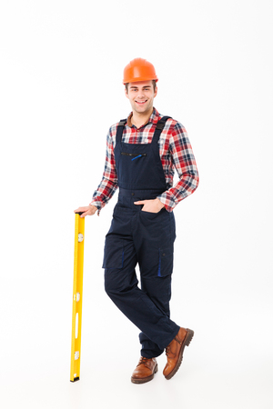 Full length portrait of a smiling young male builder holding spirit level tool while standing and looking at camera isolated over white backgroundの写真素材