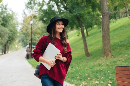Portrait of a smiling asian girl dressed in hat and sweater holding laptop computer while walking outdoorsの写真素材