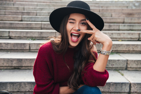 Close up portrait of a cheerful funny asian woman dressed in hat and sweater looking at camera while sitting outdoors on staircaseの写真素材