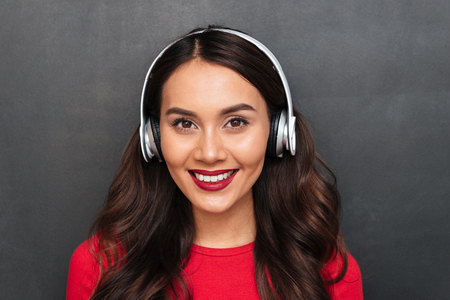Close up view of brunette woman in red blouse and headphones listening music and looking at the camera over black backgroundの写真素材