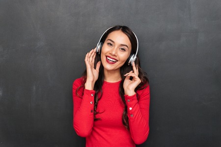 Smiling brunette woman in red blouse and headphones listening music and looking at the camera over...