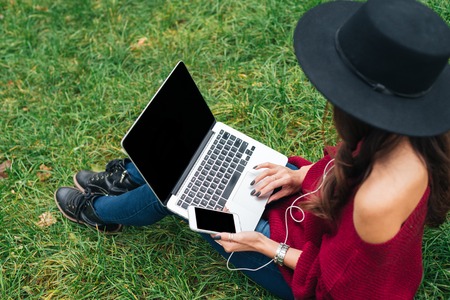 Top view of a young woman in hat using laptop computer and mobile phone while sitting on grass outdoorsの写真素材