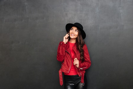 Image of happy young woman wearing hat standing over dark grey wall chalkboard talking by mobile phone. Looking aside.の写真素材