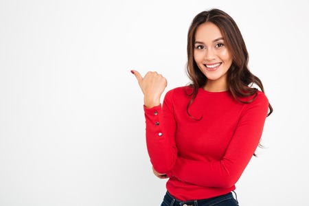 Photo of young cheerful woman standing isolated over white wall background. Looking camera pointing to copyspace.