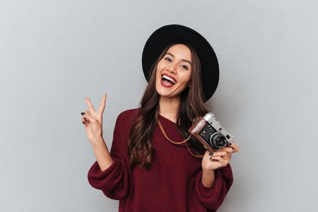 Happy brunette woman in sweater and hat holding retro camera while showing peace gesture over gray backgroundの写真素材