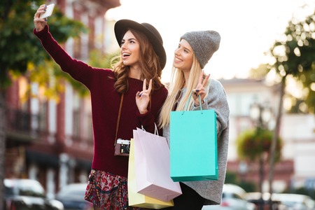 Portrait of two smiling cheerful girls holding shopping bags while standing and taking a selfie on a city streetの写真素材