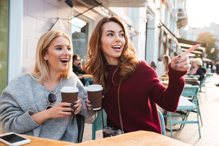 Two excited smiling girls holding coffee cups while sitting at the cafe outdoors and pointing finger awayの写真素材