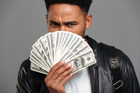 Close up portrait of a serious afro american man covering face with money banknotes isolated over...