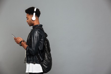 Portrait of a young afro american man in headphones using mobile phone while standing with copy...