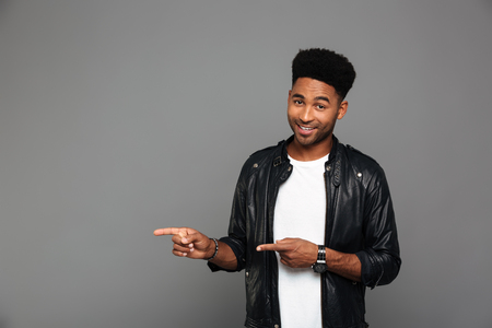 Cheerful afro american man in leather jacket pointing with two fingers, looking at camera, isolated on gray backgroundの写真素材