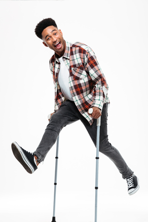 Full length portrait of a happy young afro american man jumping on crutches isolated over white backgroundの写真素材