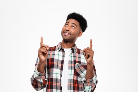 Portrait of a joyful young african man dressed in plaid shirt looking and pointing fingers up at copy space isolated over white backgroundの写真素材