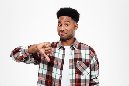 Portrait of an upset disappointed young african man dressed in plaid shirt showing thumbs down...