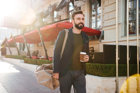 Portrait of a handsome smiling man drinking coffee while walking on a city street and carrying a bagの写真素材