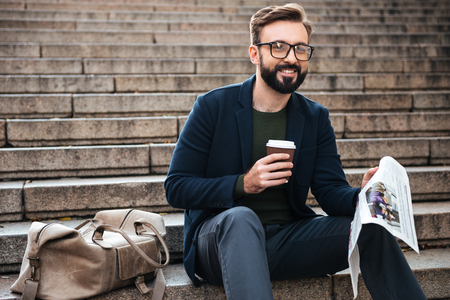 Picture of happy young bearded man sitting outdoors on steps reading newspaper and drinking coffee. Looking aside.の写真素材