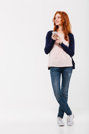 Image of happy beautiful young redhead lady standing isolated over white wall background. Looking aside chatting by mobile phone.の写真素材
